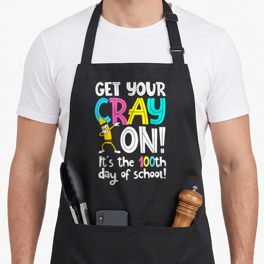 Cray teacher apron - Stacked text Get Your Cray On with dabbing yellow cartoon crayon character and 100th day of school — shown on a black cotton canvas apron against a white background