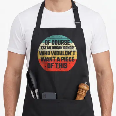 Transplant awareness organ apron - Grey text reading OF COURSE I'M AN ORGAN DONOR inside a teal beige yellow red grey — shown on a black cotton canvas apron against a white background