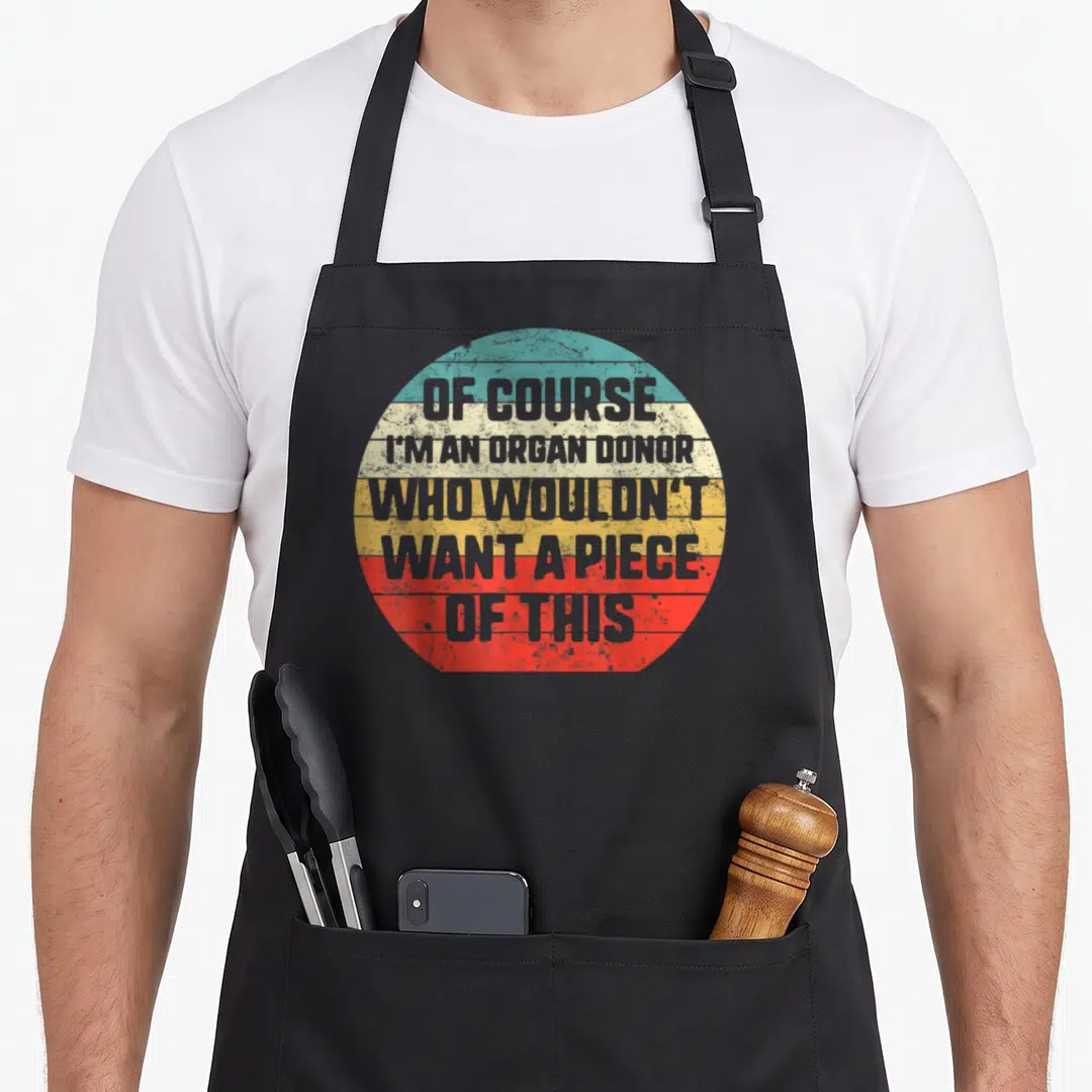 Transplant awareness organ apron - Grey text reading OF COURSE I'M AN ORGAN DONOR inside a teal beige yellow red grey — shown on a black cotton canvas apron against a white background