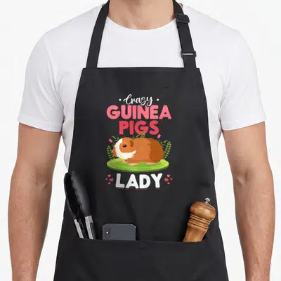 Funny guinea animal apron - Cartoon guinea pig on grass surrounded by pink text and small hearts — shown on a black cotton canvas apron against a white background