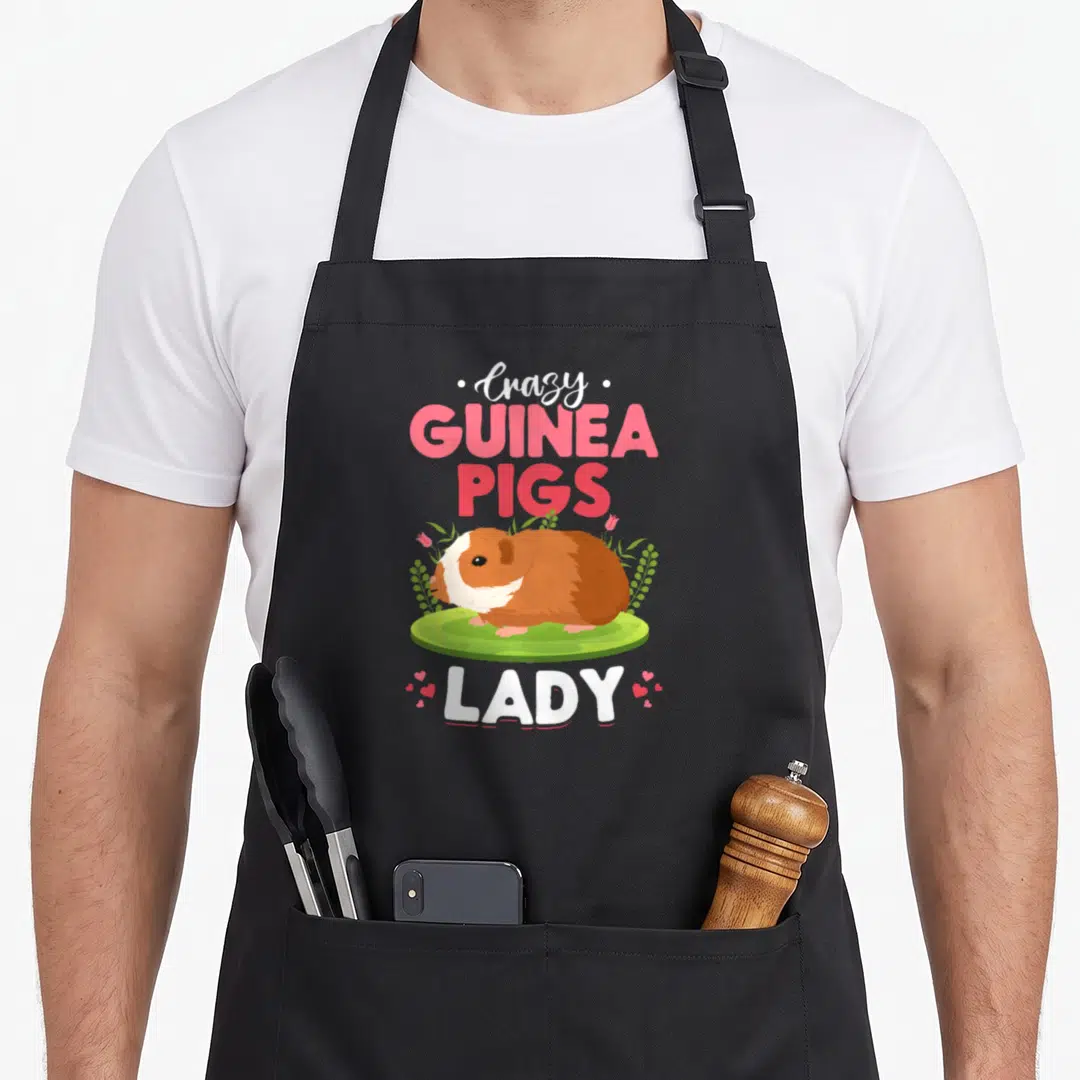 Funny guinea animal apron - Cartoon guinea pig on grass surrounded by pink text and small hearts — shown on a black cotton canvas apron against a white background