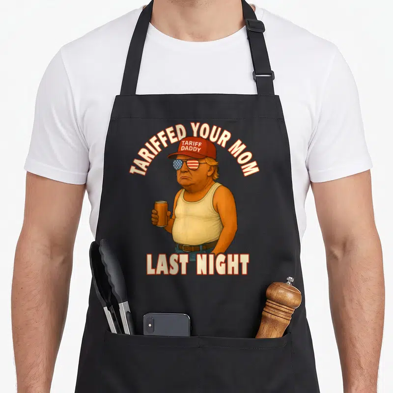 Tariffed meme apron - Cartoon man with tariff daddy hat and TARIFFED YOUR MOM LAST NIGHT text — shown on a black cotton canvas apron against a white background