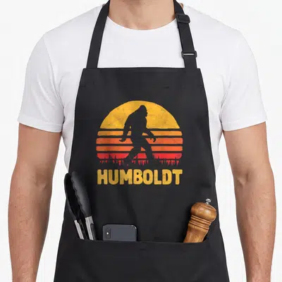 Humboldt bigfoot apron - Grey Bigfoot silhouette walking across yellow orange red striped sun with HUMBOLDT text below — shown on a black cotton canvas apron against a white background