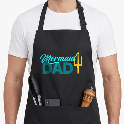 Cute mermaid birthday apron - Mermaid DAD text in blue and teal with a yellow trident symbol — shown on a black cotton canvas apron against a white background