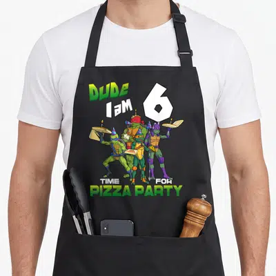 Turtle pizza apron - TMNT turtles holding pizza with DUDE I AM 6 text in green white blue purple — shown on a black cotton canvas apron against a white background