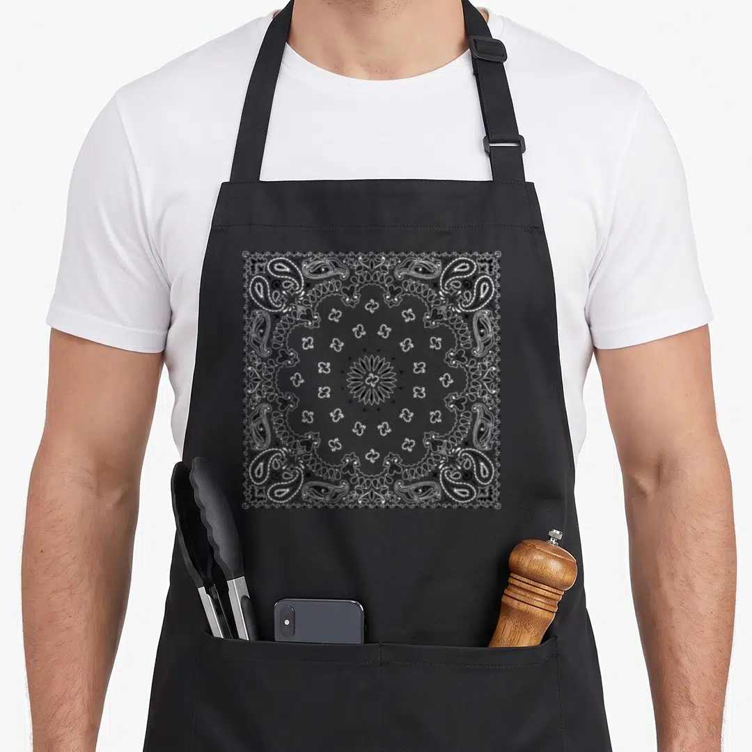 Bandana colorful apron - Grey and white symmetrical bandana pattern with central flower and radiating paisley line art — shown on a black cotton canvas apron against a white background