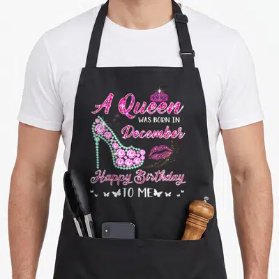 Retro december birthday apron - High heel shoe with A Queen Was Born In December text, pink gems, lips, and butterflies — shown on a black cotton canvas apron against a white background