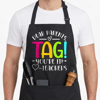 Dear parent tag apron - Stacked colorful text DEAR PARENTS TAG YOU'RE IT TEACHERS with hearts and arrow graphics — shown on a black cotton canvas apron against a white background