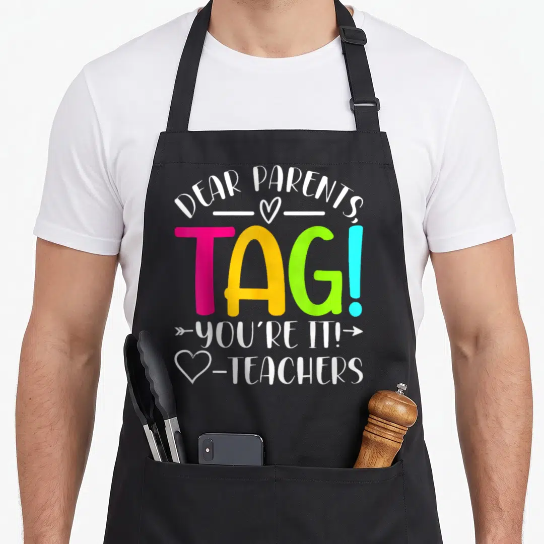 Dear parent tag apron - Stacked colorful text DEAR PARENTS TAG YOU'RE IT TEACHERS with hearts and arrow graphics — shown on a black cotton canvas apron against a white background
