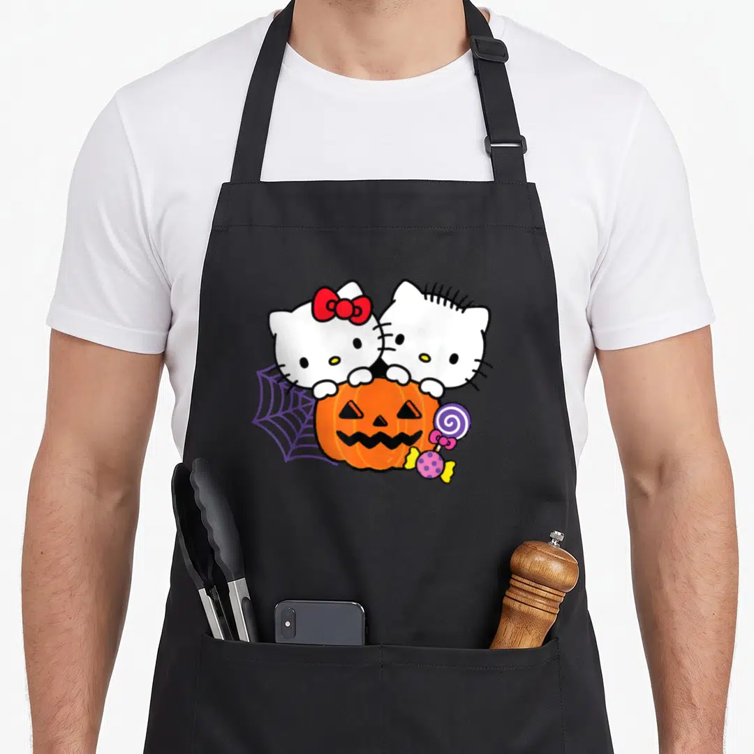 Dear halloween apron - Two white cartoon cats peeking over an orange pumpkin next to a purple spider web and lollipop — shown on a black cotton canvas apron against a white background