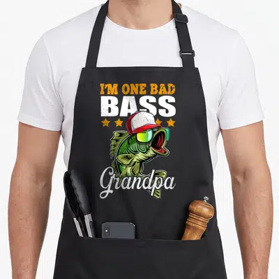 Bass fishing apron - Cartoon green bass in cap and sunglasses with I'm One Bad Bass Grandpa text — shown on a black cotton canvas apron against a white background