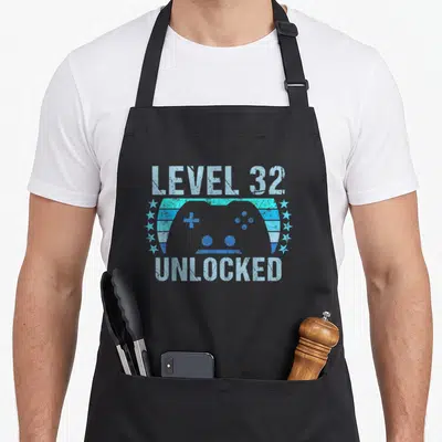 32nd gaming aprons - Distressed blue game controller between LEVEL 32 and UNLOCKED text flanked by star rows — shown on a black cotton canvas apron against a white background