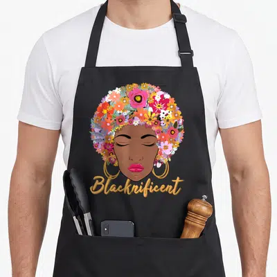 Blacknificient african american aprons - Cartoon woman face with floral afro hair in brown pink yellow red and gold — shown on a black cotton canvas apron against a white background