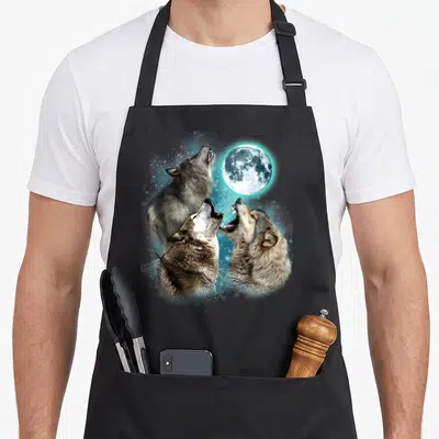 Moon wolves apron - Three realistic wolf heads howling at a full moon with blue stars — shown on a black cotton canvas apron against a white background