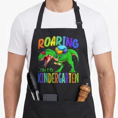 Rex school dinosaur apron - Green T-Rex with sunglasses backpack and pencil under colorful roaring into kindergarten — shown on a black cotton canvas apron against a white background