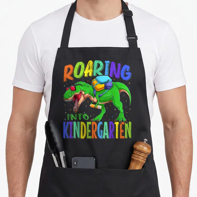Rex school dinosaur apron - Green T-Rex with sunglasses backpack and pencil under colorful roaring into kindergarten — shown on a black cotton canvas apron against a white background