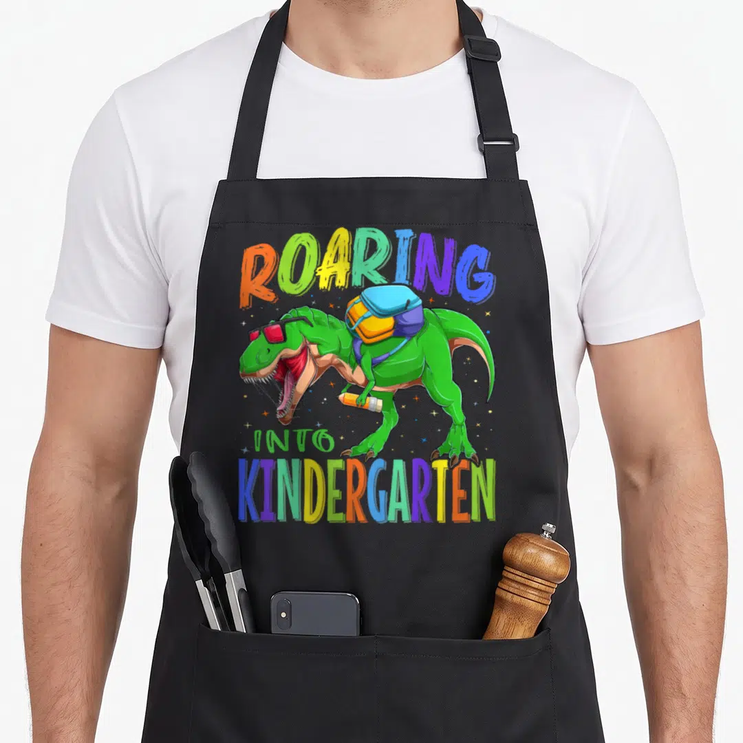 Rex school dinosaur apron - Green T-Rex with sunglasses backpack and pencil under colorful roaring into kindergarten — shown on a black cotton canvas apron against a white background