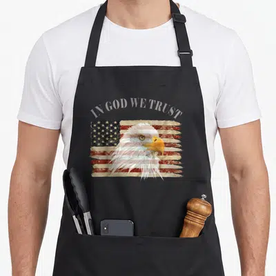 Respect trust god apron - Arched IN GOD WE TRUST text over bald eagle head on distressed flag — shown on a black cotton canvas apron against a white background
