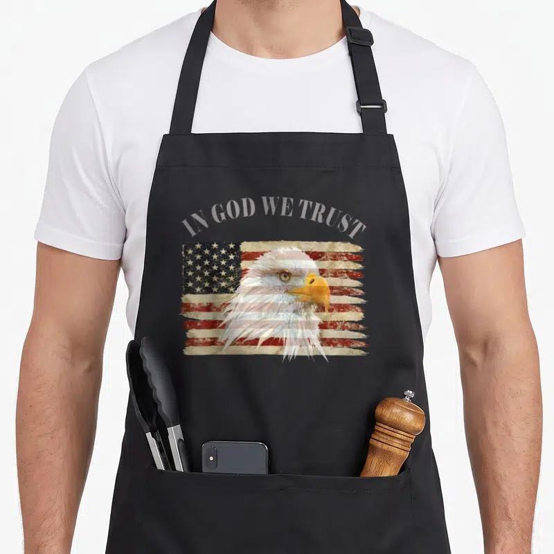Respect trust god apron - Arched IN GOD WE TRUST text over bald eagle head on distressed flag — shown on a black cotton canvas apron against a white background