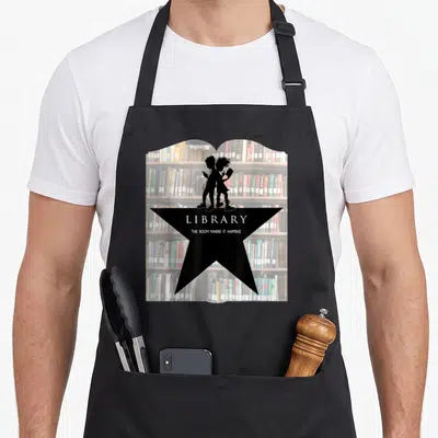 Library ny times bestseller apron - Book-shaped photo of shelves with black star, two silhouettes, and white text — shown on a black cotton canvas apron against a white background
