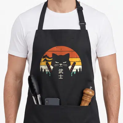 Warrior ninja cat apron - Grey cat silhouette with crossed swords and kanji text on striped sun background — shown on a black cotton canvas apron against a white background