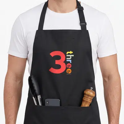 3rd age birthday apron - Red number 3 and vertical three text in yellow grey blue and orange — shown on a black cotton canvas apron against a white background