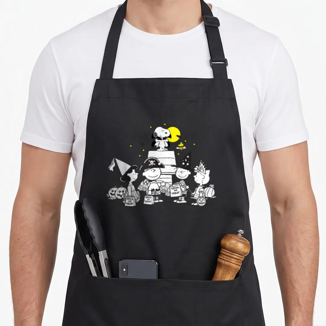 Cool halloween apron - Peanuts characters in costumes holding trick or treat bags with perched Snoopy and moon — shown on a black cotton canvas apron against a white background