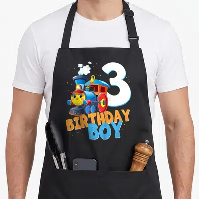 3rd train birthday apron - Cartoon blue train with red wheels next to white number 3 and BIRTHDAY BOY text — shown on a black cotton canvas apron against a white background