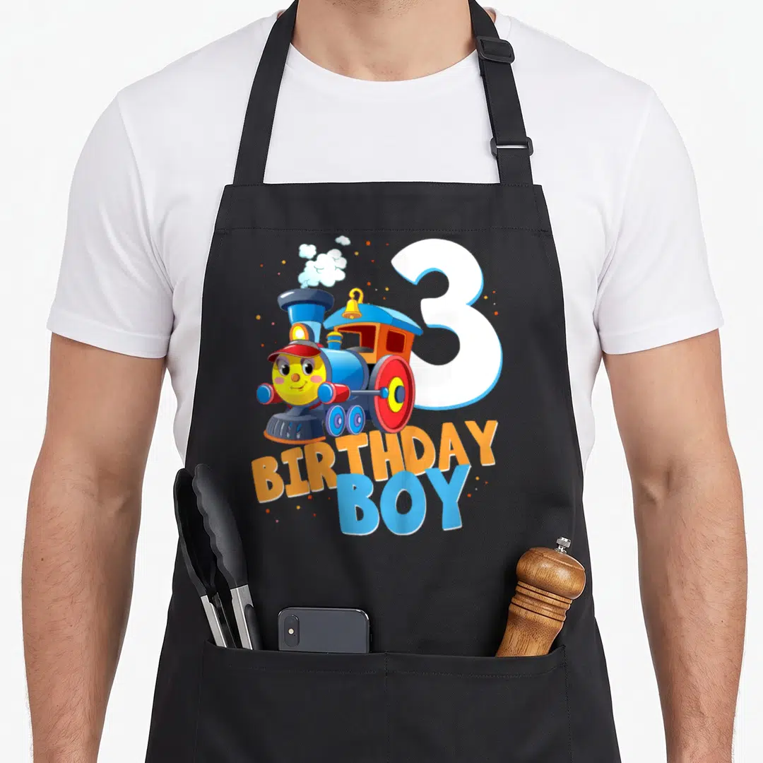 3rd train birthday apron - Cartoon blue train with red wheels next to white number 3 and BIRTHDAY BOY text — shown on a black cotton canvas apron against a white background