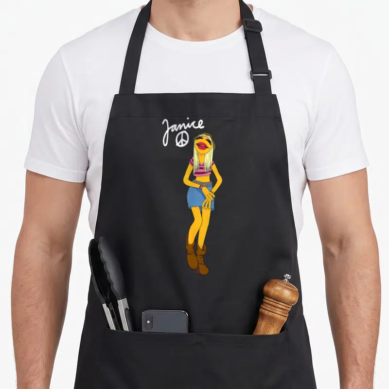 Janice big band apron - Yellow muppet wearing blue shorts and brown boots with white Janice text and peace sign — shown on a black cotton canvas apron against a white background
