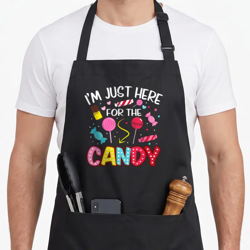 Lollipop halloween apron - Scattered lollipops and candies in white, pink, yellow, blue, red with I'M JUST HERE — shown on a black cotton canvas apron against a white background