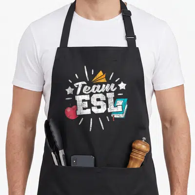 Esl school teacher apron - Distressed Team ESL text with red apple, teal book, and orange paper plane icon — shown on a black cotton canvas apron against a white background