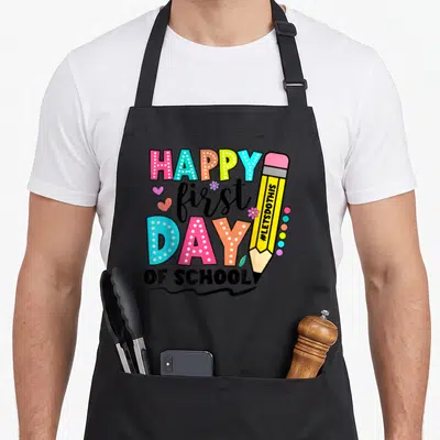 School instructor apron - Cartoon pencil and HAPPY first DAY OF SCHOOL text in pink, blue, yellow, green, and black — shown on a black cotton canvas apron against a white background