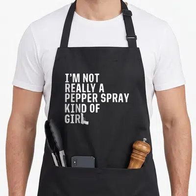 Spray pro apron - White stacked text I'm Not Really A Pepper Spray Kind Of Girl with gun silhouette replacing L — shown on a black cotton canvas apron against a white background