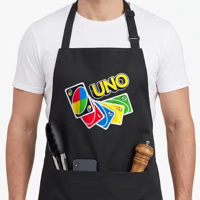 Uno game aprons - Tilted UNO logo above four fanned playing cards in yellow, red, blue, and green — shown on a black cotton canvas apron against a white background