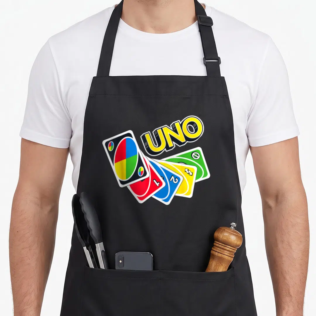 Uno game aprons - Tilted UNO logo above four fanned playing cards in yellow, red, blue, and green — shown on a black cotton canvas apron against a white background
