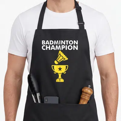 Champion badminton apron - Yellow trophy and shuttlecock below white BADMINTON CHAMPION text — shown on a black cotton canvas apron against a white background