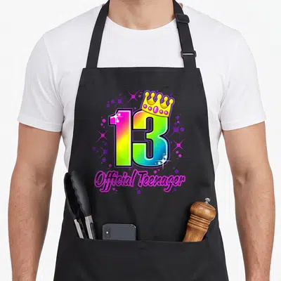 Vintage 13th general birthday apron - Rainbow number 13 with gold crown and Official Teenager text surrounded by stars — shown on a black cotton canvas apron against a white background