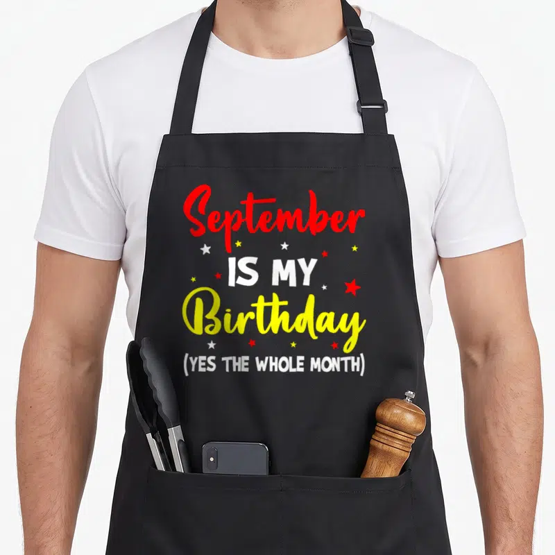 September whole month birthday apron - Stacked red yellow white text reading September IS MY Birthday YES THE WHOLE — shown on a black cotton canvas apron against a white background