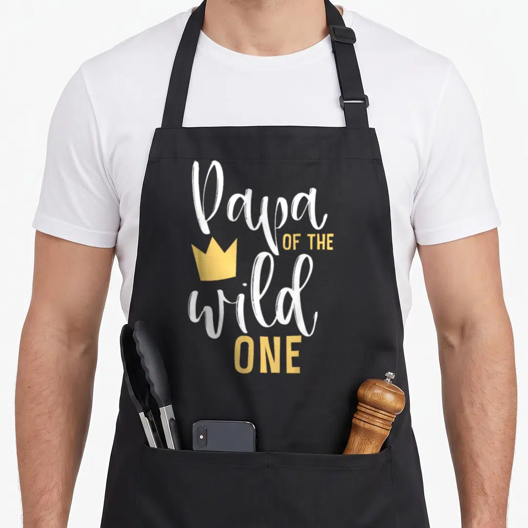 Papa wild birthday apron - Stacked white and yellow text reading Papa OF THE wild ONE with a yellow crown icon — shown on a black cotton canvas apron against a white background
