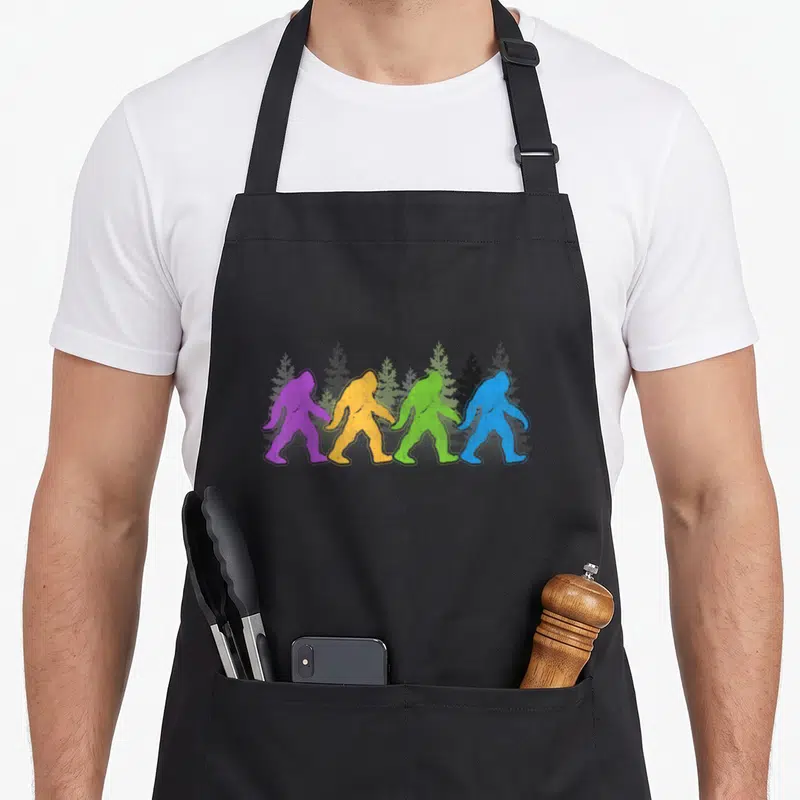 Sasquatch yeti apron - Four colorful bigfoot silhouettes walking past grey pine trees in distressed style — shown on a black cotton canvas apron against a white background