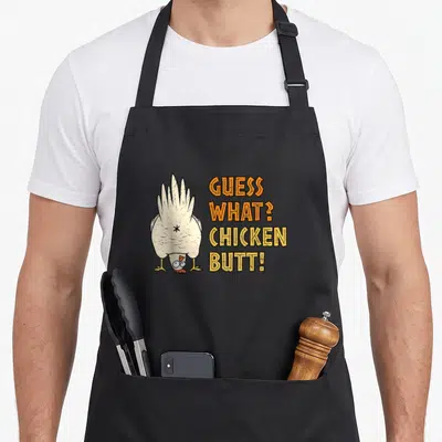 Funny guess chicken apron - Cartoon chicken viewed from behind next to stacked orange and yellow text reading GUESS — shown on a black cotton canvas apron against a white background