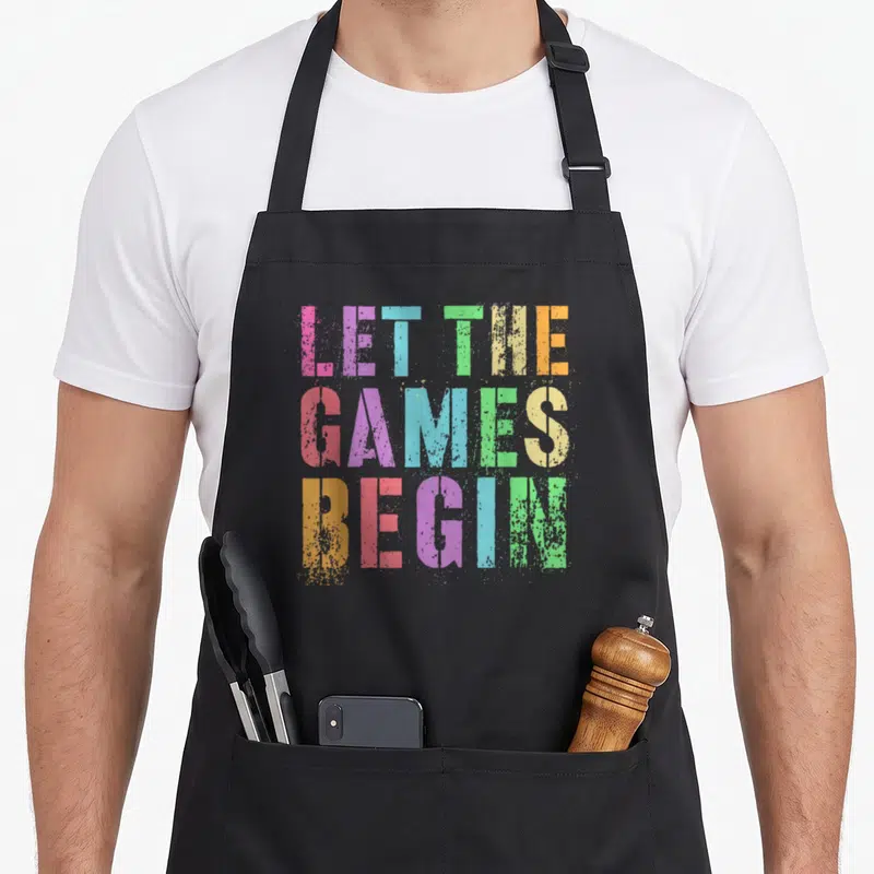 Begin game aprons - Stacked multicolor distressed text reading LET THE GAMES BEGIN on black fabric — shown on a black cotton canvas apron against a white background