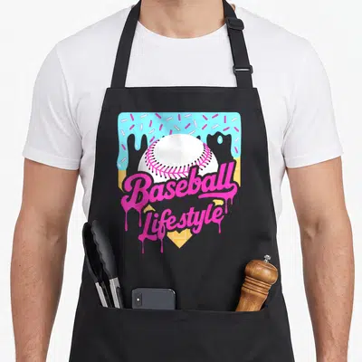 Baseball lifestyle drip apron - Pink script reading Baseball Lifestyle over white baseball with blue frosting — shown on a black cotton canvas apron against a white background