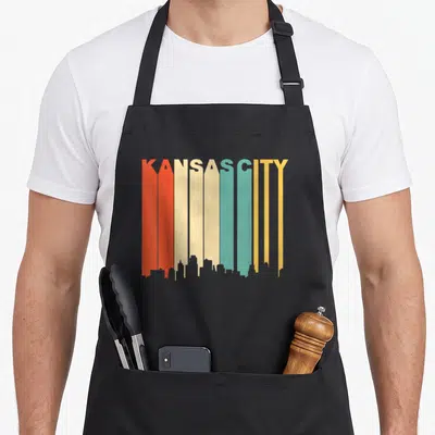 Kansas 1970s apron - KANSAS CITY text forming red orange cream teal yellow skyline bars — shown on a black cotton canvas apron against a white background
