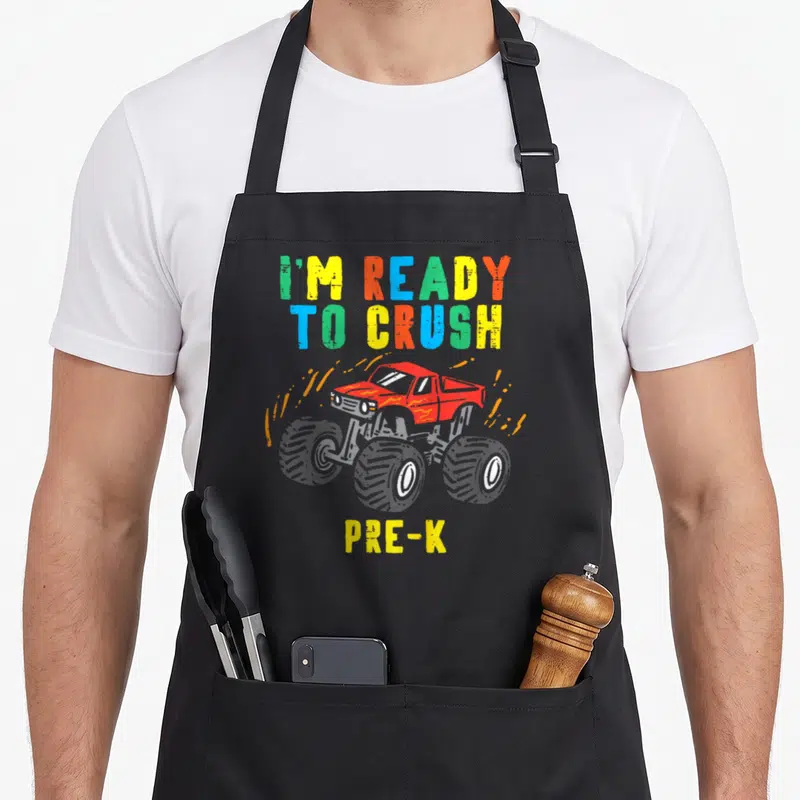 Prek truck apron - I'M READY TO CRUSH PRE-K text with red monster truck and orange dirt splashes in cartoon style — shown on a black cotton canvas apron against a white background