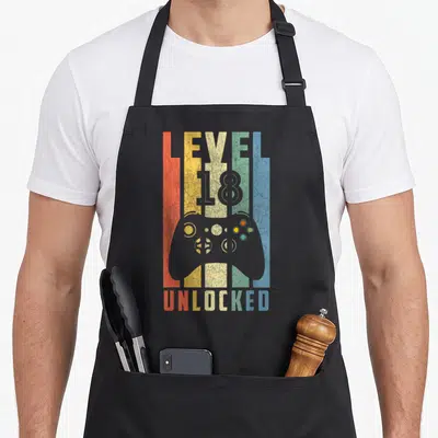 Unlocked 18th birthday apron - Retro game controller silhouette with vertical colored stripes and text reading LEVEL 18 — shown on a black cotton canvas apron against a white background