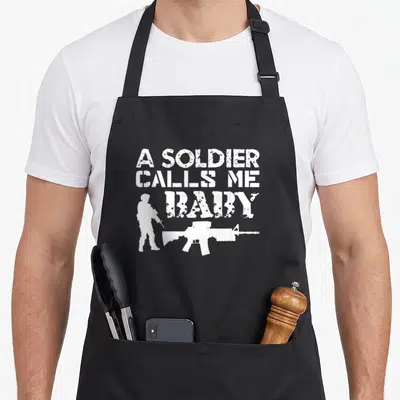 Soldier funny rings apron - White distressed text A SOLDIER CALLS ME BABY with soldier and rifle silhouettes — shown on a black cotton canvas apron against a white background