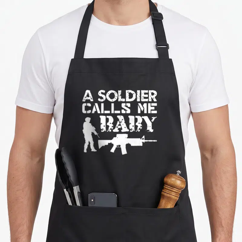 Soldier funny rings apron - White distressed text A SOLDIER CALLS ME BABY with soldier and rifle silhouettes — shown on a black cotton canvas apron against a white background