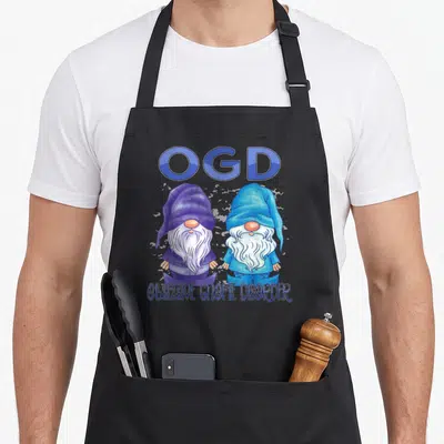 Garden gnome apron - Two cartoon gnomes in blue and purple plaid under OGD text — shown on a black cotton canvas apron against a white background